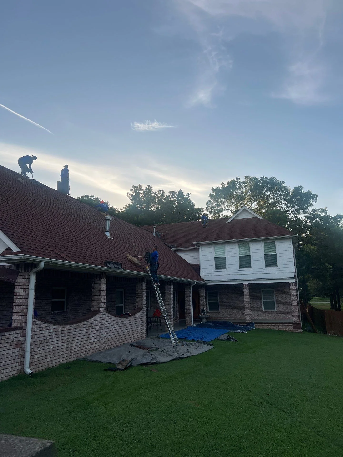 Roofers installing dark red-brown asphalt shingles on two-story brick home with protective tarps