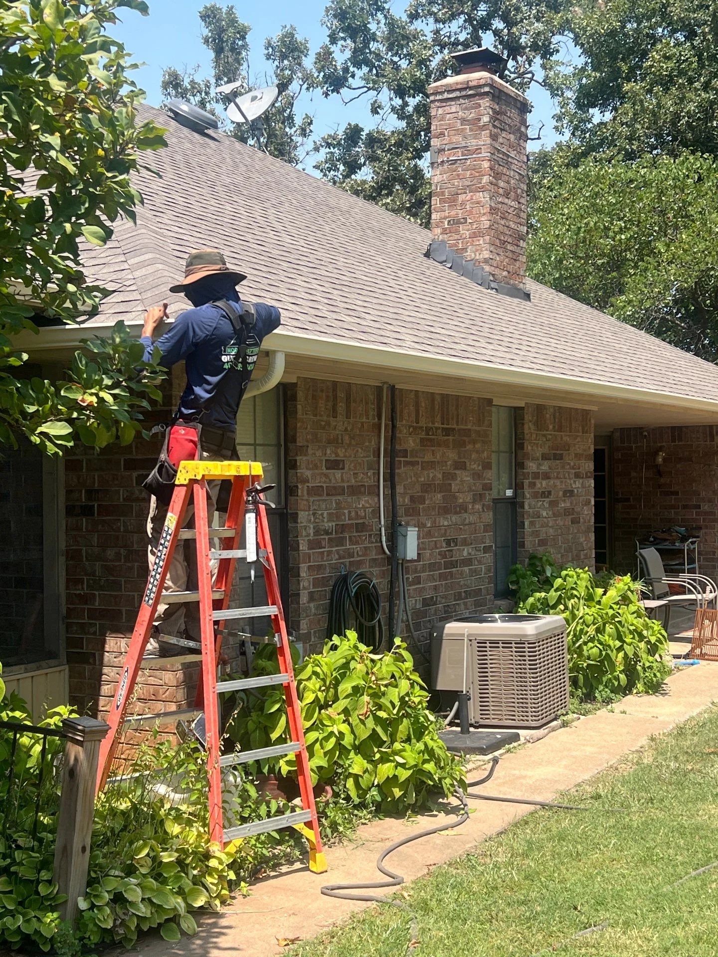 Roofer on ladder inspecting asphalt shingle roof with brick chimney on residential home